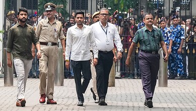 A man in a white shirt and glasses walking with police officers and other men in an outdoor public space.