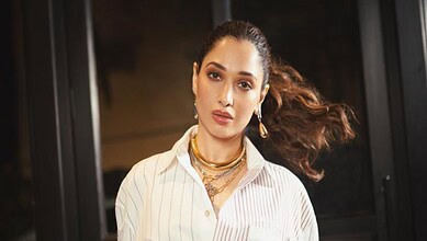 Fashionable woman with flowing hair, wearing a white striped shirt and gold jewelry, in an urban backdrop.