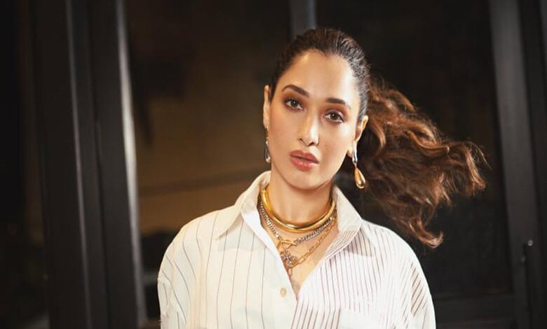 Fashionable woman with flowing hair, wearing a white striped shirt and gold jewelry, in an urban backdrop.