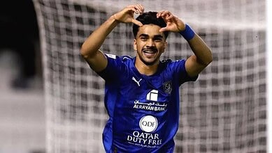 Football player celebrating goal with hands on head, smiling, wearing blue kit on the field.