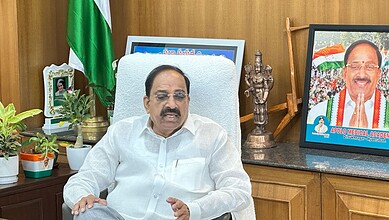 Indian politician speaking at a desk with a flag and framed photo in the background.