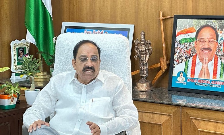 Indian politician speaking at a desk with a flag and framed photo in the background.