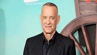 Tom Hanks at an event, wearing a black suit and shirt, smiling subtly.