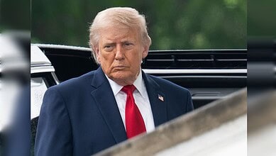 Donald Trump in a navy suit with a red tie, looking serious outdoors during a political event.