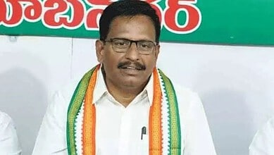 A man wearing glasses and a white shirt with a tricolour sash, standing in front of a green and white bac.