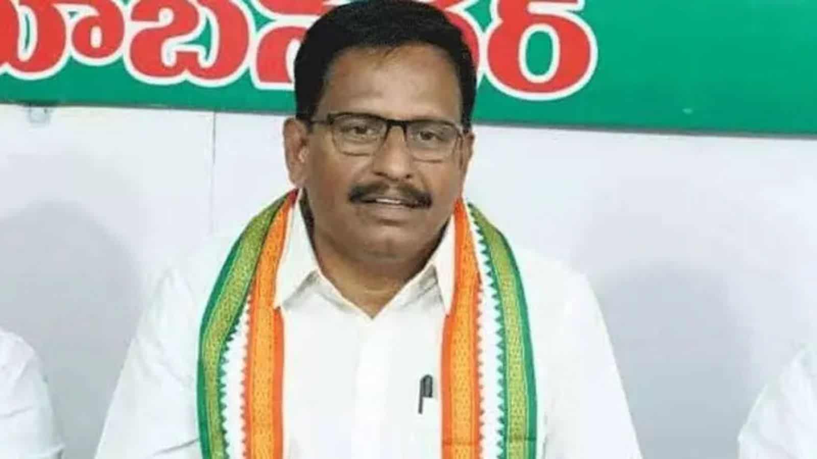 A man wearing glasses and a white shirt with a tricolour sash, standing in front of a green and white bac.