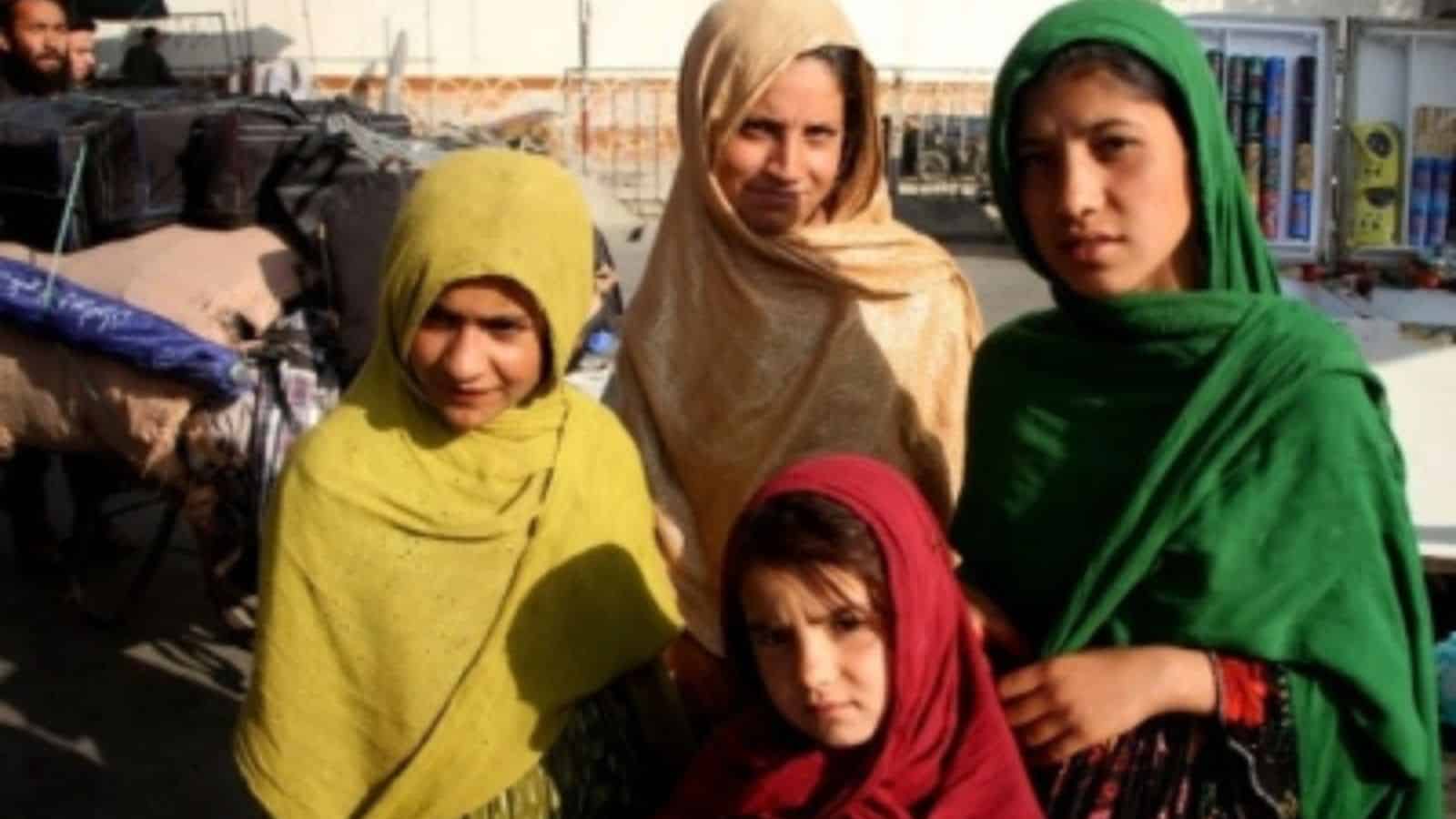 A woman and three girls wearing colorful headscarves outdoors, with a background of market stalls and bui.