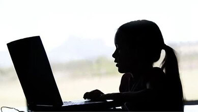 A woman using a laptop, silhouetted against a bright window, working or studying indoors.