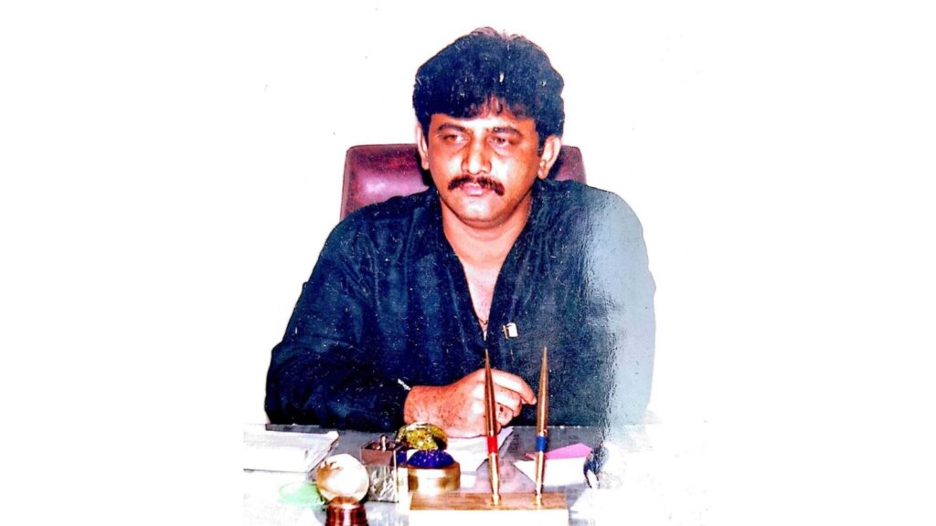 A man with dark hair and a mustache sitting at a desk with a microphone, in an office setting.