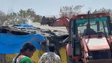 Collapsed makeshift shelter with blue tarpaulin, surrounded by trees, and a pink excavator at the site.
