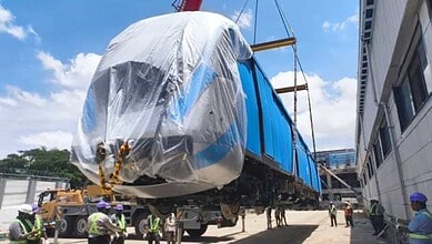 Large blue train carriage covered with plastic wrap being transported on a flatbed truck.