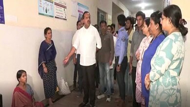 A woman sitting on the floor listening to a man speaking to a group of people in a corridor.