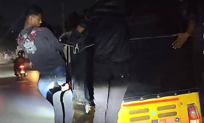 A woman and a man inspecting a black car on a street at night, with motorcycles and streetlights in the b.