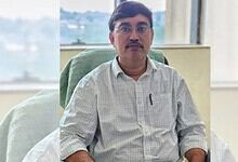 A man with glasses and a mustache sitting at a desk in an office, wearing a light checkered shirt.