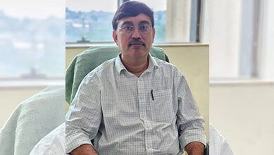 A man with glasses and a mustache sitting at a desk in an office, wearing a light checkered shirt.