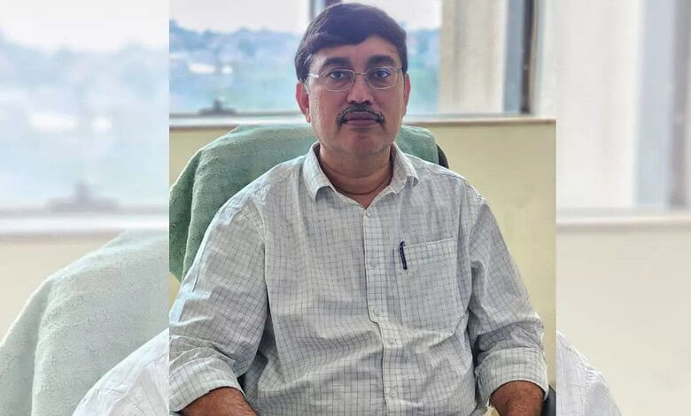 A man with glasses and a mustache sitting at a desk in an office, wearing a light checkered shirt.