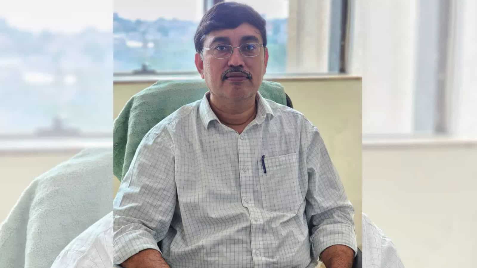 A man with glasses and a mustache sitting at a desk in an office, wearing a light checkered shirt.