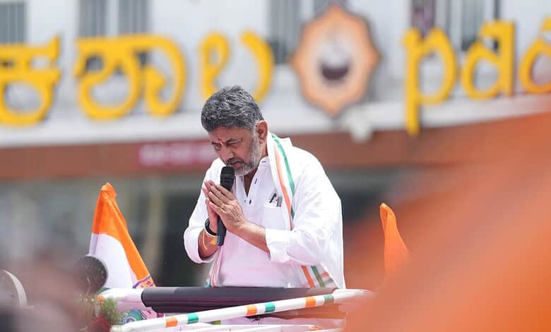 A man in white traditional attire praying with folded hands in front of a microphone during a public even.