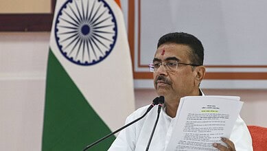 Indian politician speaking at a podium with the Indian flag in the background, wearing glasses and tradit.