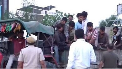 A man selling fruits from a cart with a group of young people gathered around, outdoors in an urban setti.