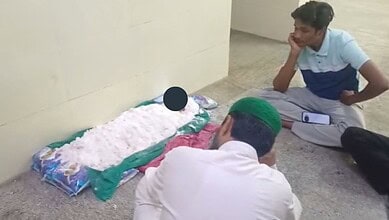 A young man praying on the floor with a green cap, facing a makeshift prayer area with a pillow and white.