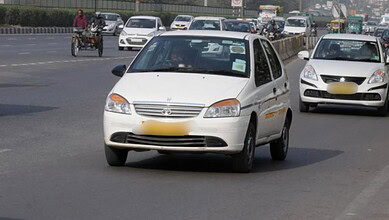 A white compact car driving on a busy city road with multiple vehicles and motorcycles in the background.