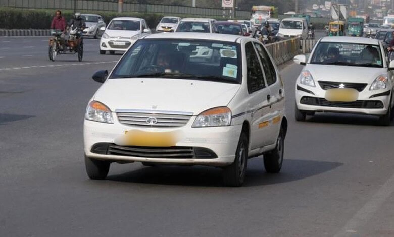 A white compact car driving on a busy city road with multiple vehicles and motorcycles in the background.