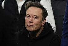 Elon Musk speaking at a public event, wearing a black hoodie, with a focused expression on his face.