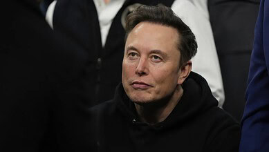 Elon Musk speaking at a public event, wearing a black hoodie, with a focused expression on his face.