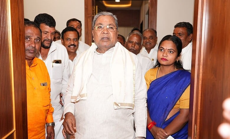 Senior man in white traditional attire with a group of people, including a woman in a blue saree, standin.