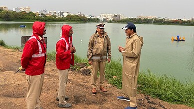 Group of four men in outdoor jackets and hats talking by a river with city buildings in the background.