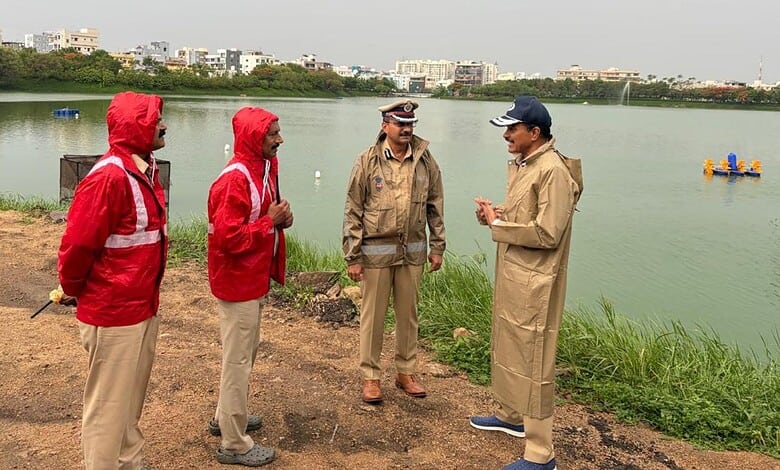 Group of four men in outdoor jackets and hats talking by a river with city buildings in the background.