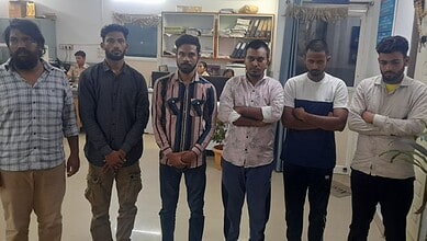 A group of six young men standing indoors in a casual setting, some with arms crossed, in front of booksh.