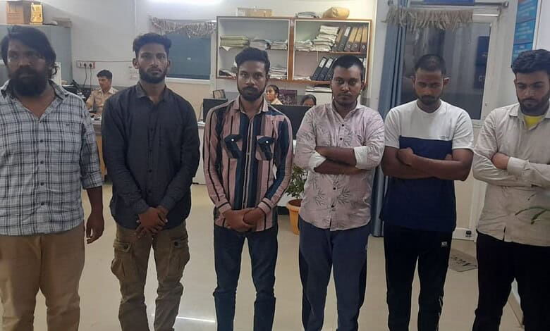 A group of six young men standing indoors in a casual setting, some with arms crossed, in front of booksh.