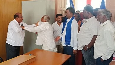 A man in a white shirt greets another man in a white kurta with a handshake in front of a group of people.