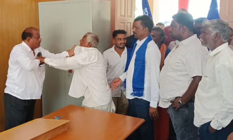 A man in a white shirt greets another man in a white kurta with a handshake in front of a group of people.