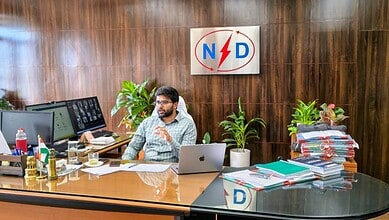 The image shows a man working at a cluttered desk in an office with a wooden wall background and a sign w.
