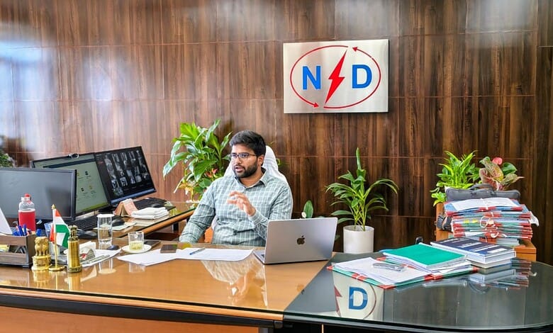 The image shows a man working at a cluttered desk in an office with a wooden wall background and a sign w.