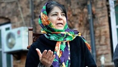A woman wearing a colorful headscarf speaking outdoors, with a brick wall and air conditioning unit in th.