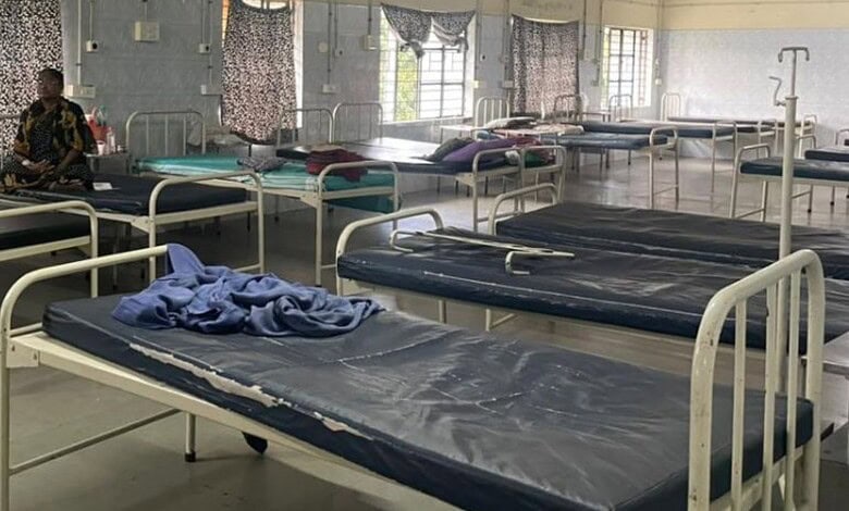 Empty hospital beds in a ward with a person sitting in the background, under natural light.