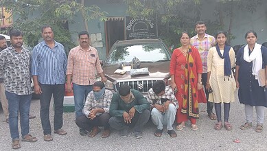 Group of nine people, including men and women, standing and sitting in front of a car outdoors.
