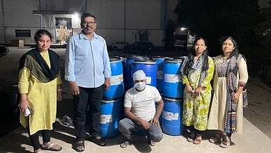 Group of five people standing outdoors near blue barrels at night, some wearing masks.