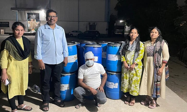Group of five people standing outdoors near blue barrels at night, some wearing masks.