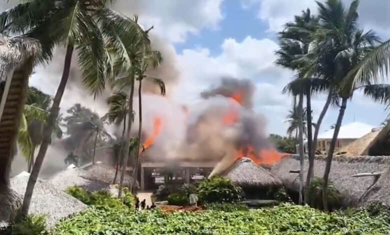 Resort on fire with thick smoke and flames, surrounded by palm trees in a tropical setting.