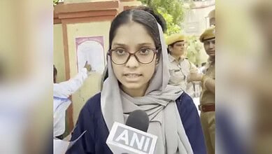Young woman in glasses and hijab speaking to media outside exam centre with police officers nearby.