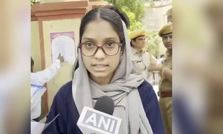 Young woman in glasses and hijab speaking to media outside exam centre with police officers nearby.