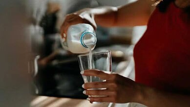Person pouring milk into a glass, focus on hand and glass, kitchen background.