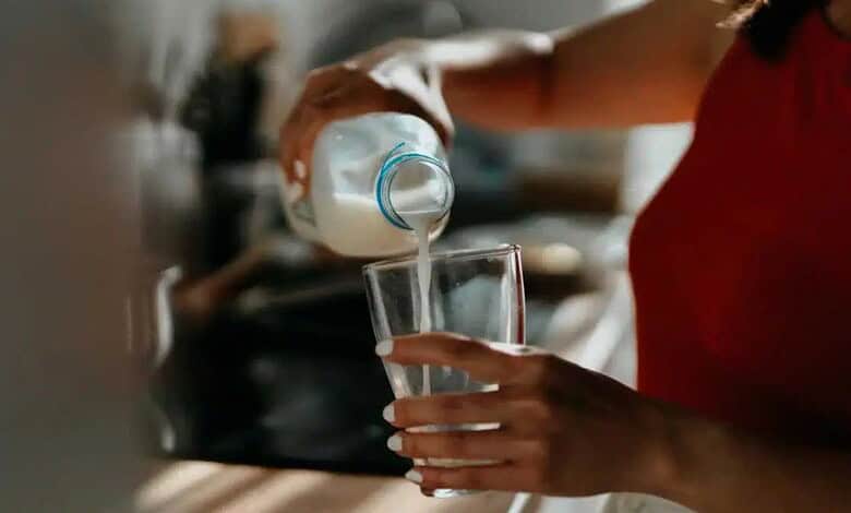 Person pouring milk into a glass, focus on hand and glass, kitchen background.