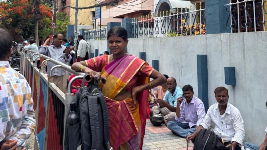Parents and students outside a school, some sitting on the ground, waiting during NEET exam day.