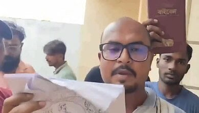 Man holding a book with others in the background, indoors, serious expression.